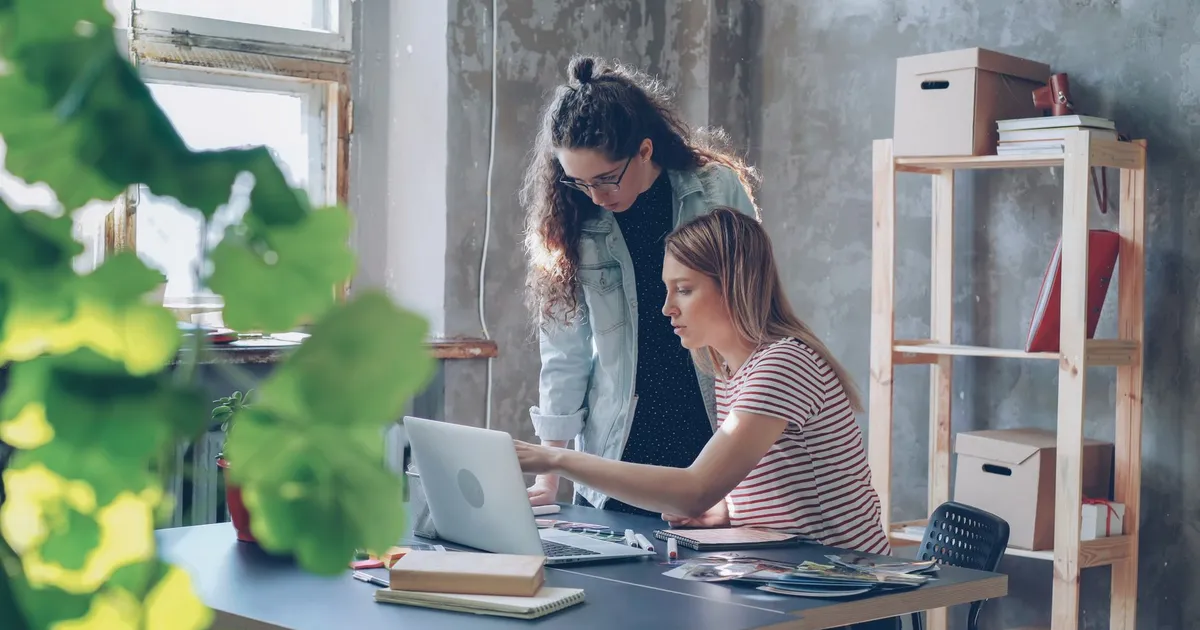 Two young women working together in a modern office with a laptop and plants.