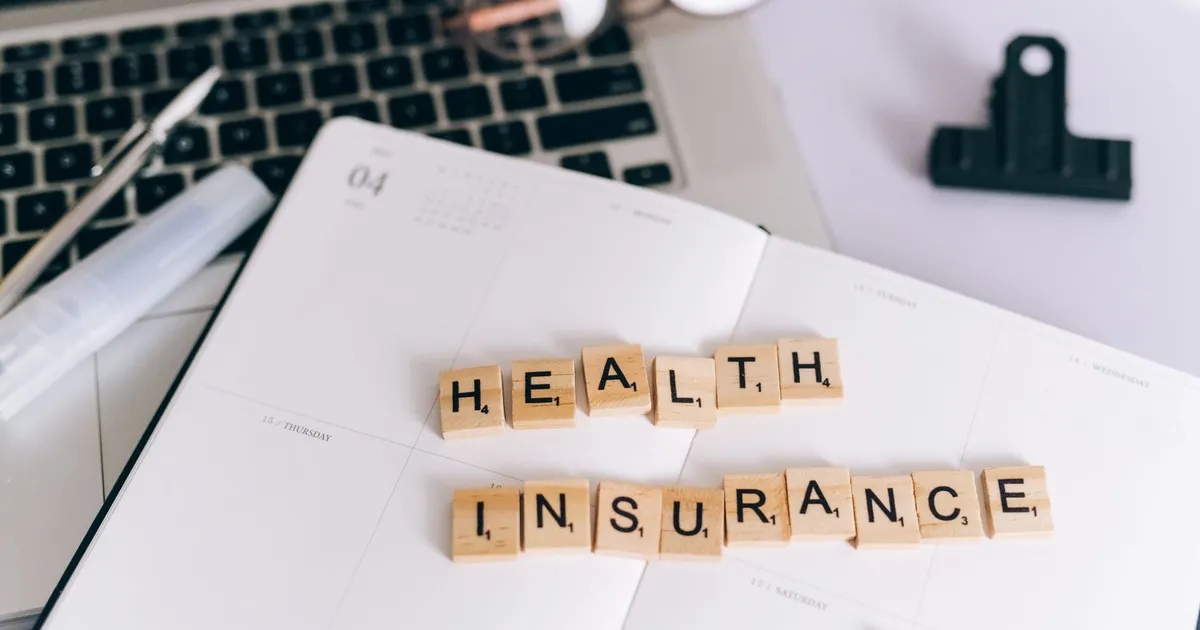 Close-up of Scrabble tiles spelling 'Health Insurance' on a planner with a laptop.