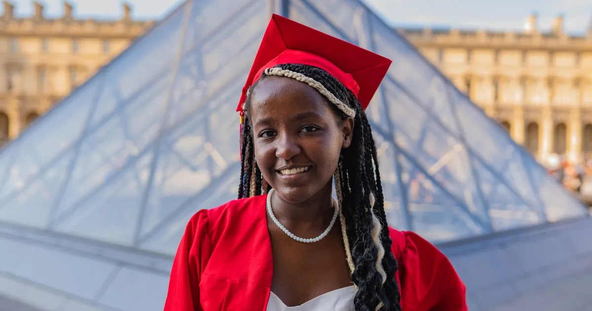 Young woman in graduation attire smiling at Louvre Pyramid, Paris.