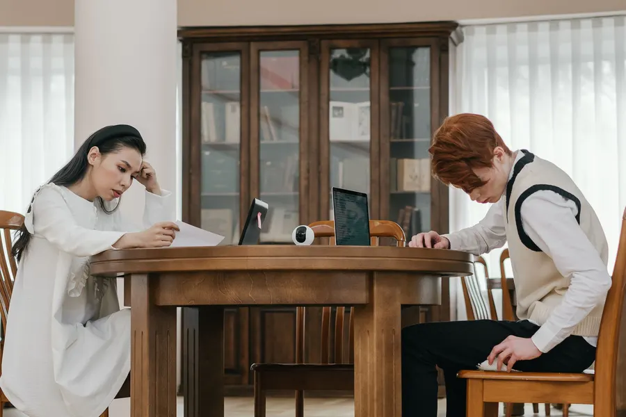 Two people studying at a wooden table with laptops and books indoors.