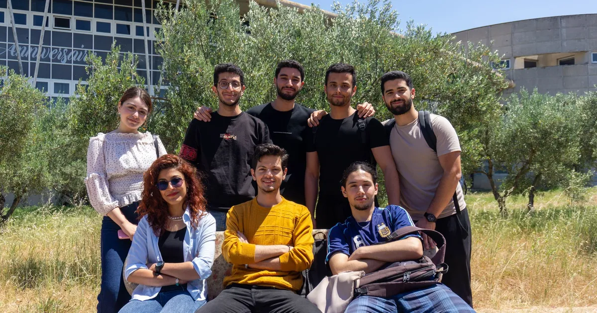 A diverse group of young adults posing together with smiles on a sunny day outdoors. University campus setting.