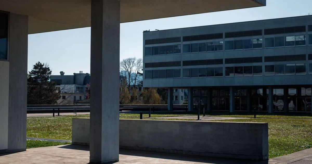 A modern concrete university building alongside an open, green field on a sunny day.