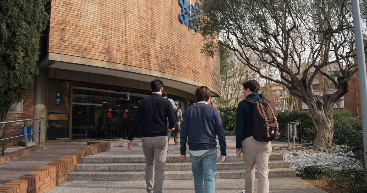 Three students approach a university building entrance in Barcelona, Spain on a sunny day.