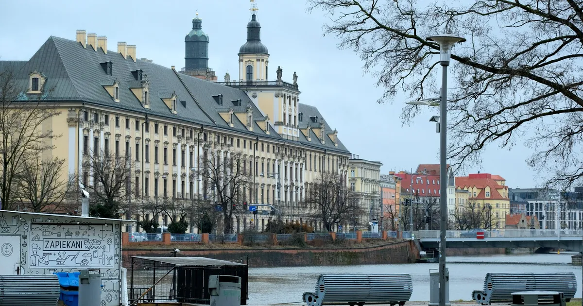 Scenic view of a historic university building by the river in Wrocław on an overcast day.
