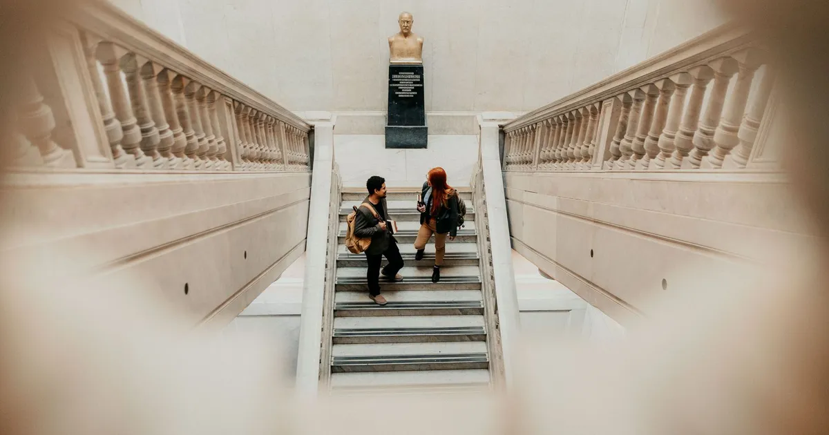 Two students talking on a grand staircase with a bust in Buenos Aires university.