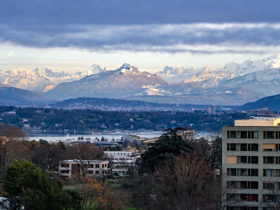 A breathtaking view of Geneva with Swiss Alps backdrop under a cloudy sky at day.
