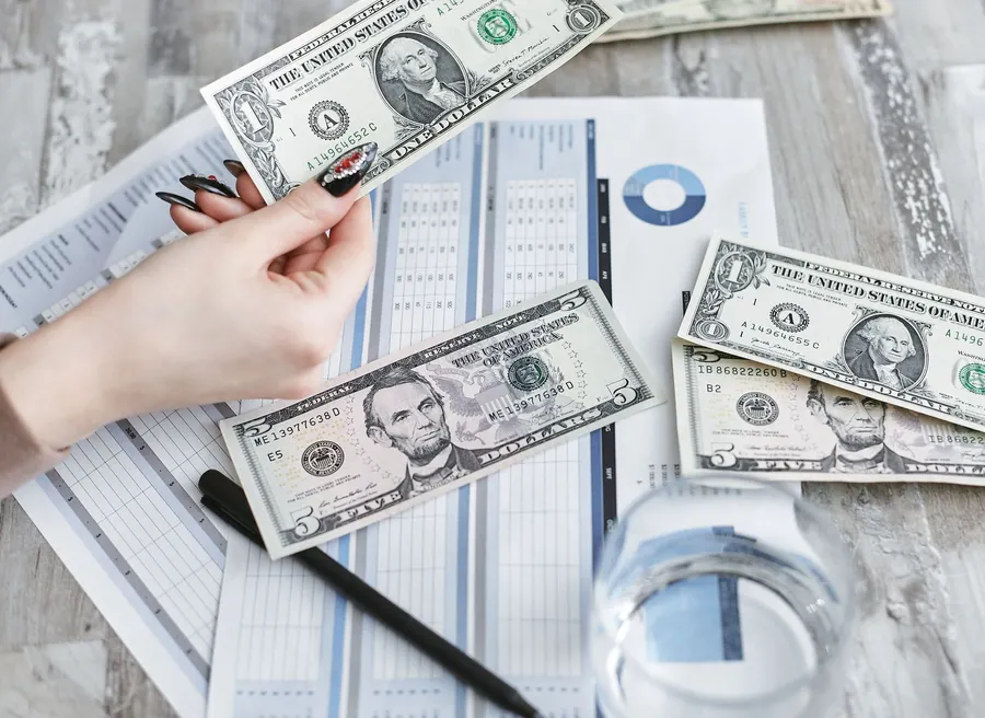 Close-up of a hand holding a dollar bill over financial documents and charts on a wooden table.