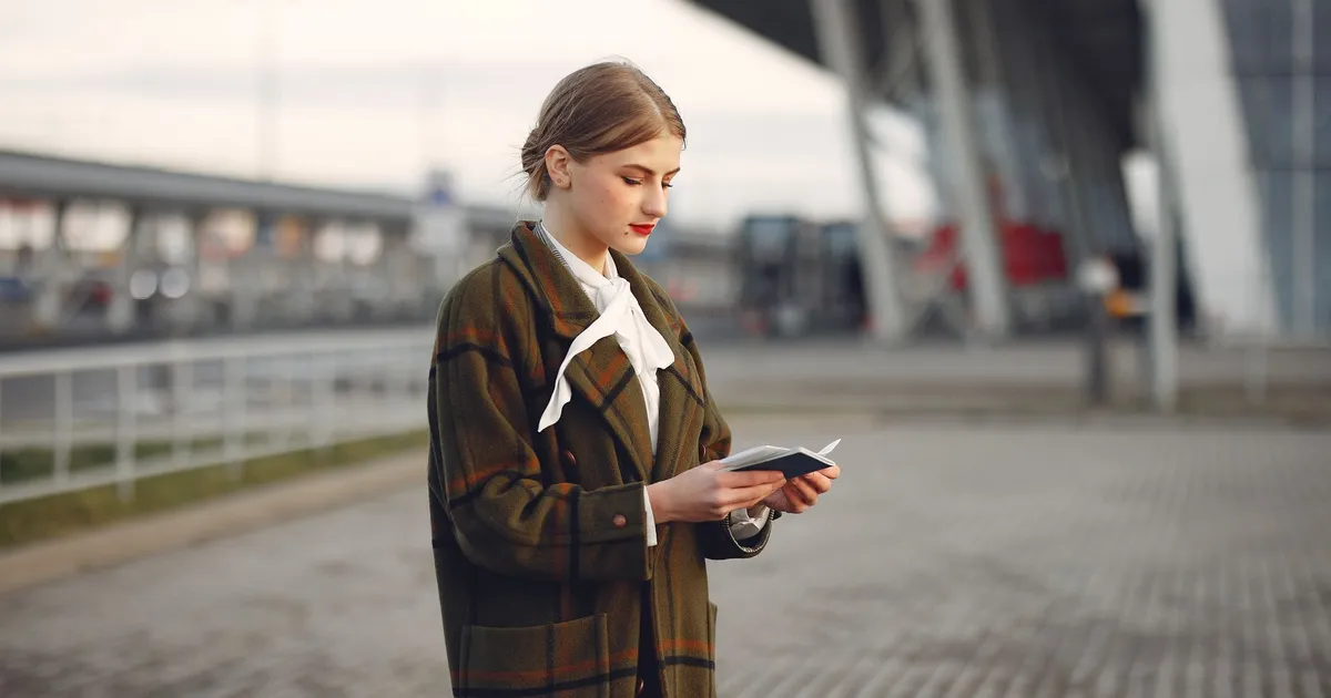 Attentive female passenger wearing trendy plaid coat and white blouse checking passport and ticket standing on pavement near modern building of airport outside