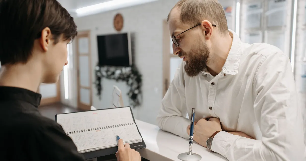 A receptionist and client converse over an appointment book at a clinic reception desk.